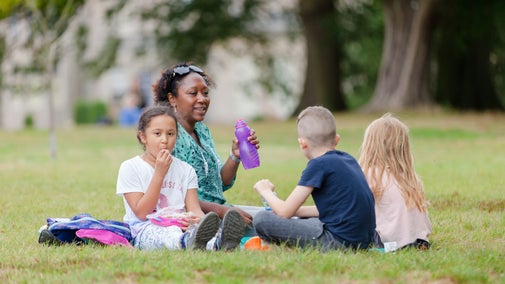 Four people sit together on a grassy lawn in a park, having a picnic. One person holds a purple drinks bottle while a child in the foreground eats from a lunchbox. Two other children sit nearby with their backs partly to the camera. Trees and buildings are softly blurred in the background, and bags and picnic items are placed on the grass.